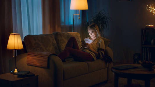 温暖房间在晚上,美丽的年轻女人躺在沙发上,使用智能手机