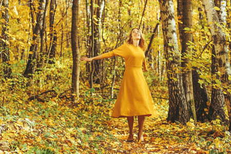 年轻漂亮的女孩穿着黄色的长裙，带着落叶在秋天的公园散步。