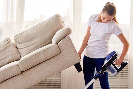 白色衬衫和牛仔裤的年轻女人的肖像，沙发下的清洁地毯，客厅里有吸尘器，复印空间。家务清洁和家务概念