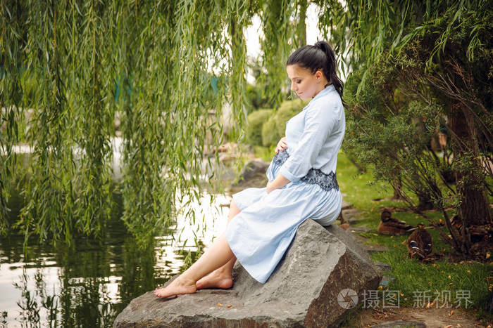 年轻美丽的孕妇放松在岩石上的湖上的背景在夏天