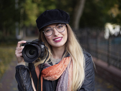 秋天公园里，一位美丽时尚开朗年轻的金发摄影师戴着眼镜，戴着一顶带妆的帽子，手里拿着一相机。