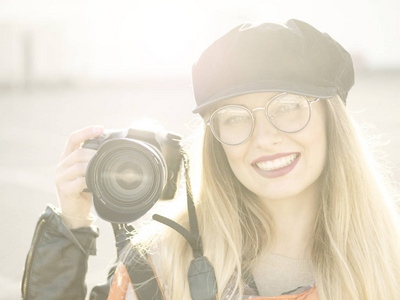 一位漂亮时尚开朗的年轻金发摄影师戴着眼镜和一顶带着黑色相机的帽子，戴着一个浅色背景的饼干。