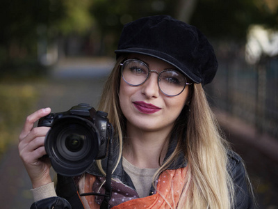 秋天公园里，一位美丽时尚开朗年轻的金发摄影师戴着眼镜，戴着一顶带妆的帽子，手里拿着一相机。