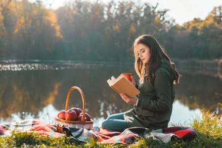 美丽的年轻女子，在公园池塘附近的毯子上放着咖啡杯看书