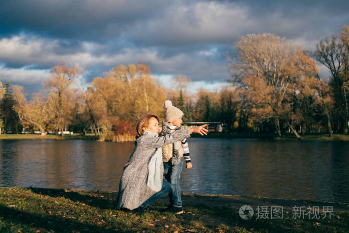 母亲和儿子坐在秋天公园的湖边日落时看着远方一家人