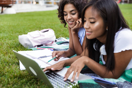 照片快乐微笑的年轻多民族朋友女孩做作业使用笔记本电脑户外公园。