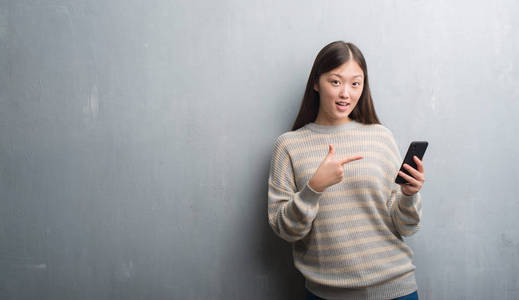 年轻的中国女人在灰色的墙壁上看着智能手机，非常高兴地用手和手指指点