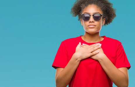 年轻的美国黑人妇女戴着太阳镜在孤立的背景上微笑，双手放在胸部，闭着眼睛，脸上有感激的手势。 健康概念。