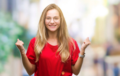 年轻漂亮的金发女人戴着眼镜在孤立的背景上庆祝惊讶和惊讶的成功，手臂抬起和睁开眼睛。 赢家概念。