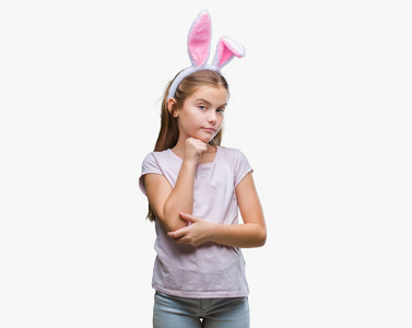 年轻漂亮的女孩戴着复活节兔子耳朵在孤立的背景上，手在下巴上思考问题，沉思的表情。 带着体贴的脸微笑。 怀疑概念。