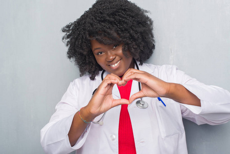 年轻的非裔美国医生女人在灰色的格墙上微笑，在爱中显示出心脏的象征和形状与手。 浪漫的概念。