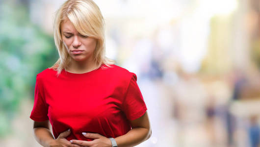 年轻漂亮的金发女人穿着红色T恤，在孤立的背景上，手放在肚子上，因为恶心疼痛疾病感觉不适。 疼痛的概念。