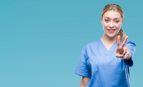 年轻的金发外科医生，女医生，穿着医疗制服，在孤立的背景上微笑，快乐的脸对镜头眨眼，做胜利标志。 二号。