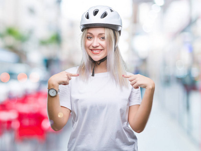 年轻的金发女人戴着自行车安全头盔，在孤立的背景下，自信地微笑着，用手指指着自己，骄傲和快乐。