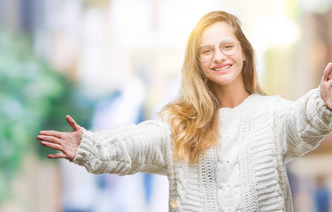 年轻漂亮的金发女人穿着冬天的毛衣和太阳镜，在孤立的背景上看着相机，张开双臂微笑着拥抱。 拥抱幸福的快乐表情。