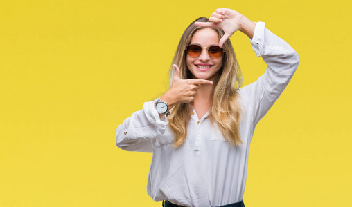 年轻漂亮的金发女人戴着太阳镜，在孤立的背景上微笑，用手和手指制作框架，用快乐的脸。 创造力和摄影理念。