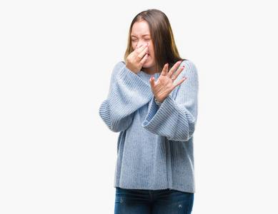 年轻美丽的白种人女人穿着冬天的毛衣，在孤立的背景上闻着一些臭味和令人恶心的难以忍受的气味，用手指在鼻子上屏住呼吸。 难闻的概念。