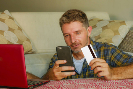 年轻英俊快乐的年轻人拿着信用卡坐在家里的沙发上，在互联网商业和电子商务概念中使用笔记本电脑和手机在线购物
