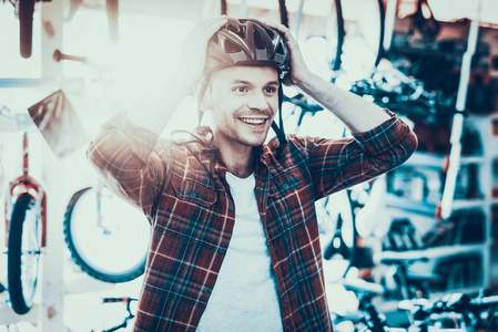 英俊快乐的家伙试着戴上头盔骑自行车。 快乐微笑的白种人顾客的肖像，穿着红色格子衬衫，在运动自行车商店里测量现代头盔，背部模糊