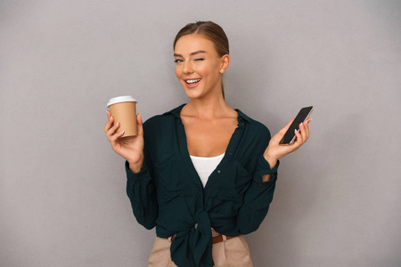 兴奋的美丽商务女性的形象，摆出孤立的灰色墙壁背景，喝咖啡使用手机。