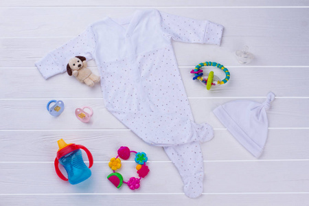 婴儿衣服和木材上的属性。白色睡衣，结帽，奶嘴，响尾蛇，瓶子和毛绒狗。木制桌子背景。