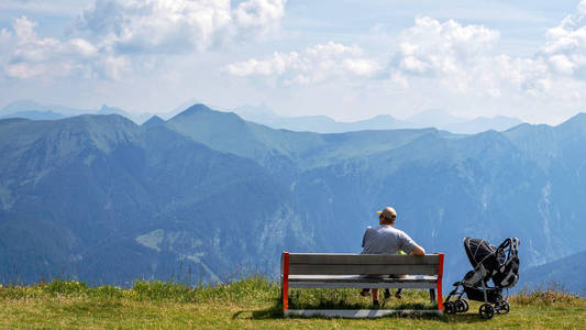 一个带着孩子的男人坐在阿尔卑斯山顶的木凳上，看着美丽的山景。