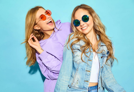 两个年轻漂亮的金发微笑的时髦女孩穿着时髦的夏装。 无忧无虑的女人戴着太阳镜在蓝色墙壁附近摆姿势。 积极的模特疯狂拥抱