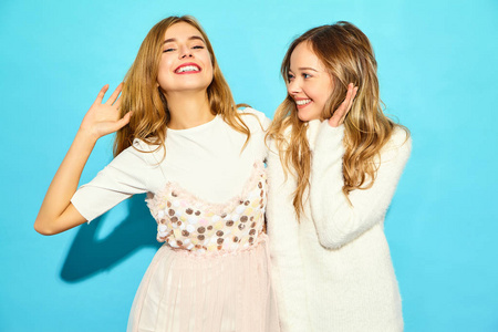 两个年轻美丽微笑的时髦女孩穿着时髦的夏季白色衣服。 无忧无虑的女人在蓝墙附近摆姿势。 正面模型