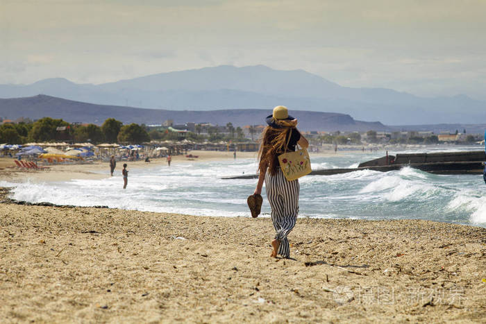 那个留着长发的女孩正在希腊的海滩上散步. 海滩上戴帽子的女人.
