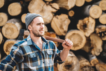 在锯木厂和树木仓库的背景下，身穿格子衬衫的强壮的长胡子伐木工人手里拿着斧头