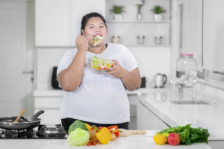 年轻胖女人站在厨房里吃一碗美味的沙拉的照片