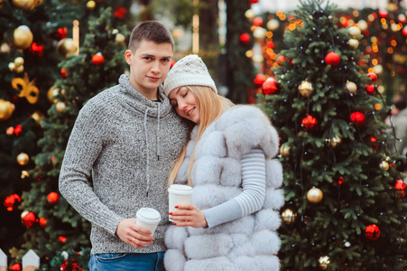 圣诞老人的肖像画，这对幸福的夫妇带着热覆盖的葡萄酒或茶走在为节日装饰的城市街道上，背景上有灯光和玩具的圣诞树