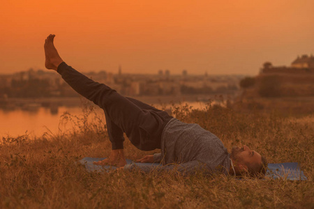 在日落时做瑜伽的人与城市景观一腿桥姿势EKAPADASetubandhaSarvangasana.toned图像。