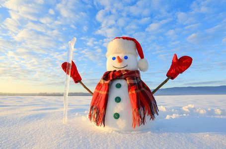 难以置信的日出。戴着帽子围巾的快乐雪人戴着冰皮的红手套站在雪地上。山景背景上..