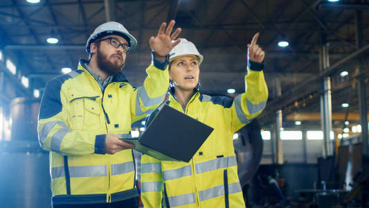 男女工业工程师在重工业工厂行走时讨论并使用笔记本电脑。他们穿硬帽和安全夹克。