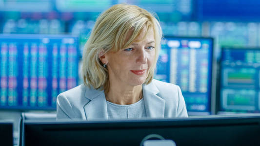 在工作站工作的资深女性股票交易员的肖像。在她的多个监视器后面显示数据滴答数字和图形。