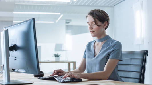 美丽的创意女性在光明现代办公室的电脑上工作。她有短发和穿着宇宙灰色连衣裙。
