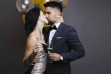 英俊的男人和可爱的女人穿着优雅的服装，戴着香槟酒杯，热情地接吻，站在灰色背景的气球附近。