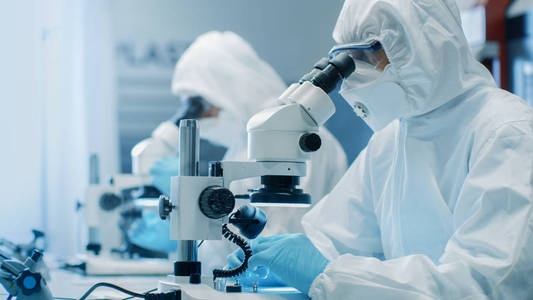 无菌洁净室套装的两名工程师科学家技术人员使用显微镜进行部件调整和研究。 他们在一家电子元器件制造厂工作。
