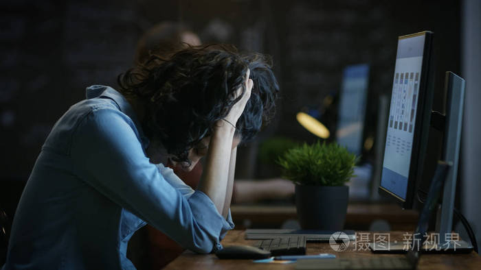 劳累过度的女性手机应用程序设计师在个人电脑上工作时，双手抱着她的头。 在后台创意办公室。