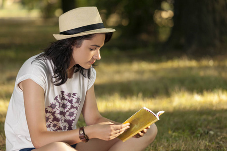 年轻漂亮的女孩，戴着帽子，坐在大自然的草地上，仔细地读着嘘声
