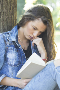 穿着牛仔裤夹克的漂亮女孩，一个学生坐在树旁边的草地上看书。 她在大自然中放松，享受一个美丽而阳光明媚的春天夏天