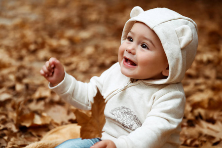 一个可爱的小男孩的肖像，手里拿着一片树叶，坐在地上，上面覆盖着秋天公园里干燥的树叶，快乐的童年概念