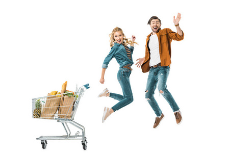 一对幸福的年轻夫妇在购物车旁跳着，里面装满了白色纸袋中的产品