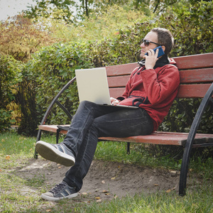 在户外工作的人总是连接到他的智能手机和他忠实的笔记本电脑。年轻人, 自由职业者在秋天公园户外工作