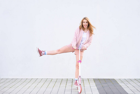 一个穿着粉红色衣服的有趣女孩在白色墙壁的背景上骑着一辆粉红色的踢踏板，并感到高兴。 疯狂时尚的女孩骑着一辆白色背景的滑板车。 共