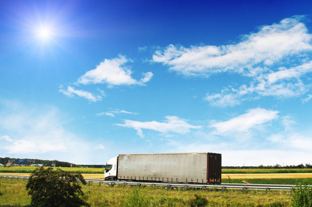 蓝色的卡车，拖车在乡间的道路上，田野和绿树在蓝天上，云彩和太阳
