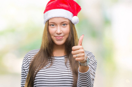 年轻美丽的高加索女人戴着圣诞帽，在孤立的背景下，用手做快乐的竖起大拇指的手势。 赞许的表情看着相机，显示出成功。