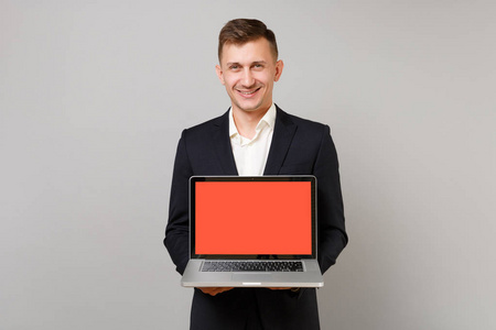 微笑的年轻商人穿着经典的黑色西装衬衫，手提电脑电脑，空白的空屏幕隔离在灰色背景上。 成就职业财富商业理念。 模拟复制空间