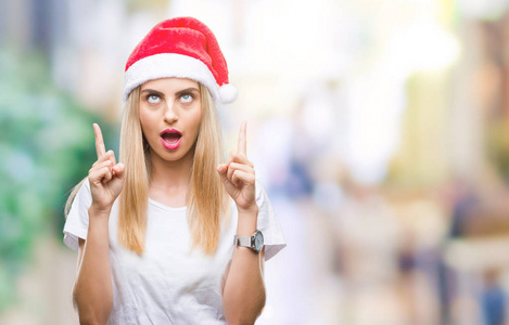 年轻漂亮的金发女人圣诞帽在孤立的背景上惊讶和惊讶地抬头，手指和举起的手臂。